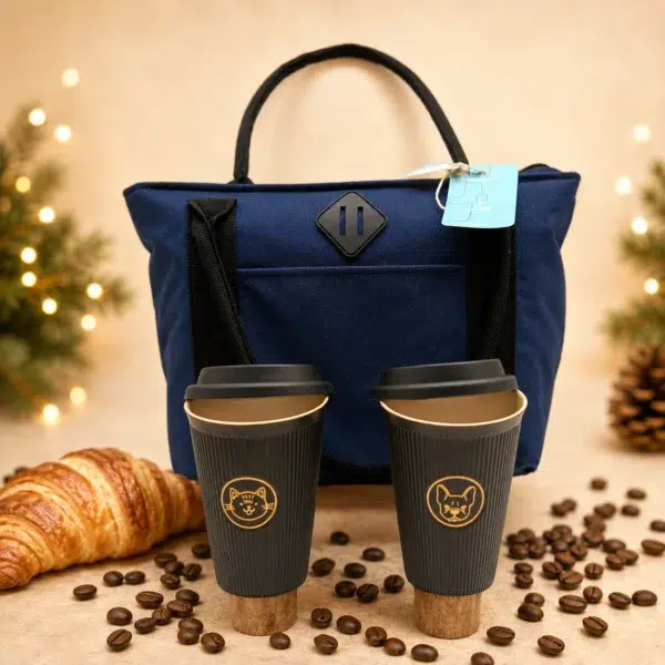 Sac isotherme bleu accompagné de deux mugs nomades noirs, d’un croissant et de grains de café dans une ambiance festive.